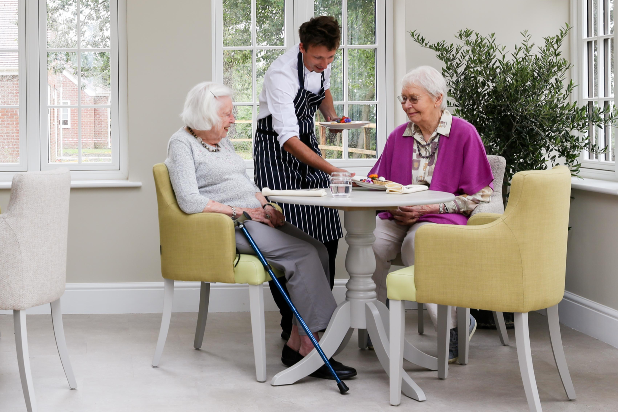 A person serves food to an elderly woman at a table
